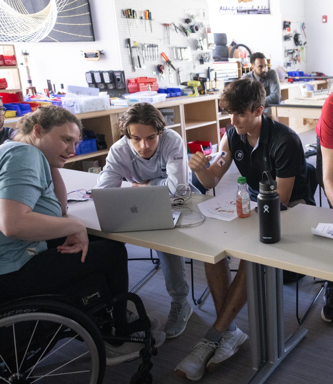 High school students and an adaptive athlete in a wheelchair working together at a laptop in a design workshop
